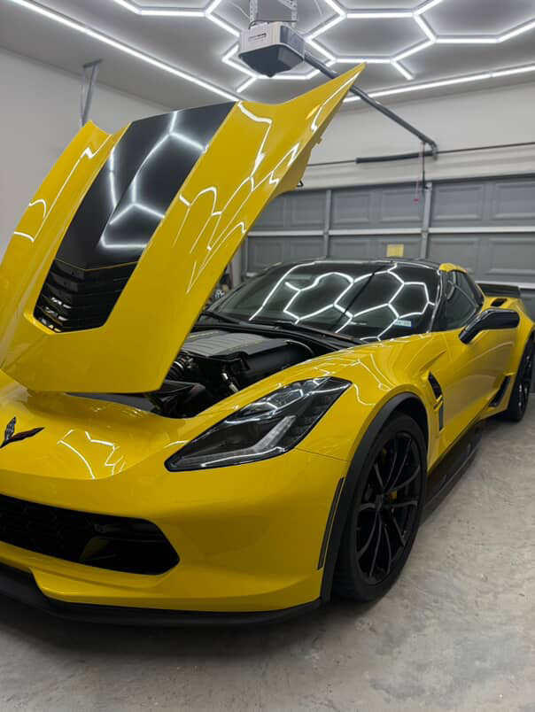 A bright yellow C7 Corvette Z06 in a detailing bay with the hood open, hexagonal LED ceiling lights reflecting across the glossy paint. A bright yellow C7 Corvette Z06 in a detailing bay with the hood open, hexagonal LED ceiling lights reflecting across the glossy paint.