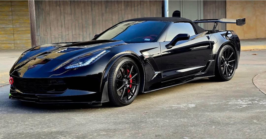A black C7 Corvette Z06 convertible with a carbon-fiber rear wing, aggressive aero, and black wheels with red calipers, parked outside a wooden building. A black C7 Corvette Z06 convertible with a carbon-fiber rear wing, aggressive aero, and black wheels with red calipers, parked outside a wooden building.