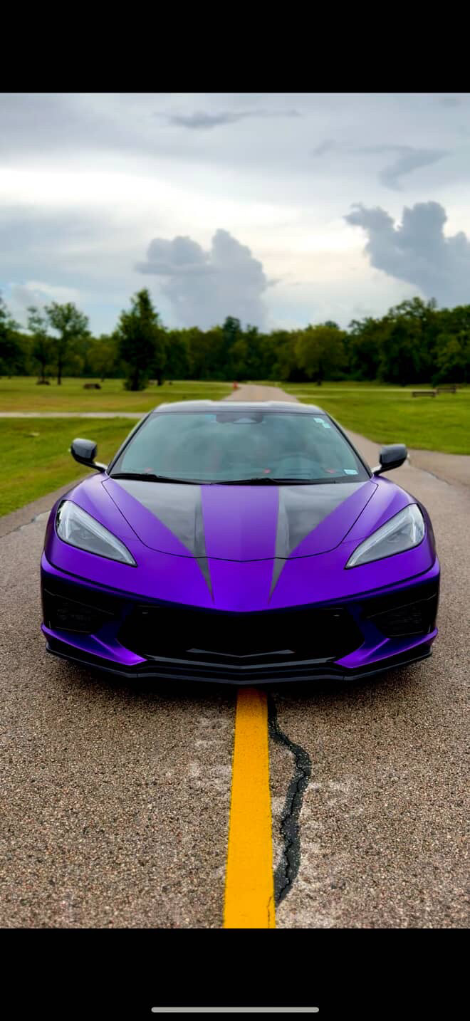 A custom purple C8 Corvette with dark grey racing stripes down the hood, parked on a rural road surrounded by green trees. A custom purple C8 Corvette with dark grey racing stripes down the hood, parked on a rural road surrounded by green trees.
