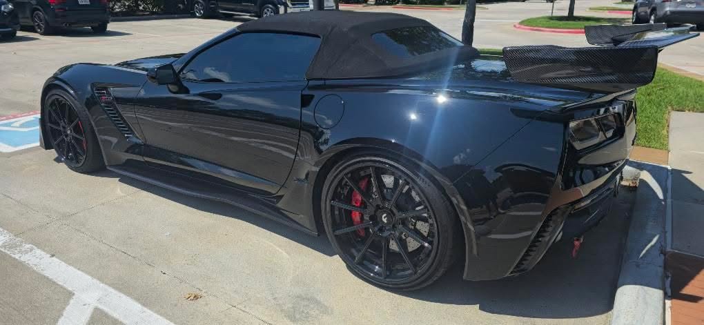 A black C7 Corvette convertible with a large carbon-fiber rear wing and black wheels with red calipers, parked in a sunlit lot. A black C7 Corvette convertible with a large carbon-fiber rear wing and black wheels with red calipers, parked in a sunlit lot.
