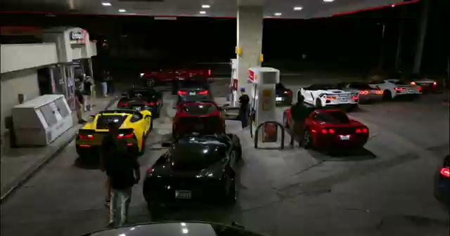 A nighttime gas station stop during an HTX Corvettes cruise — yellow C7, dark sports car, and red C7 queued at the pumps with members standing around. A nighttime gas station stop during an HTX Corvettes cruise — yellow C7, dark sports car, and red C7 queued at the pumps with members standing around.