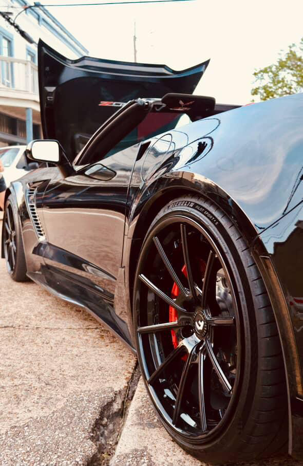A low-angle shot of a black C7 Corvette Z06 with its hood open, showcasing a gloss-black aftermarket wheel and red caliper. A low-angle shot of a black C7 Corvette Z06 with its hood open, showcasing a gloss-black aftermarket wheel and red caliper.